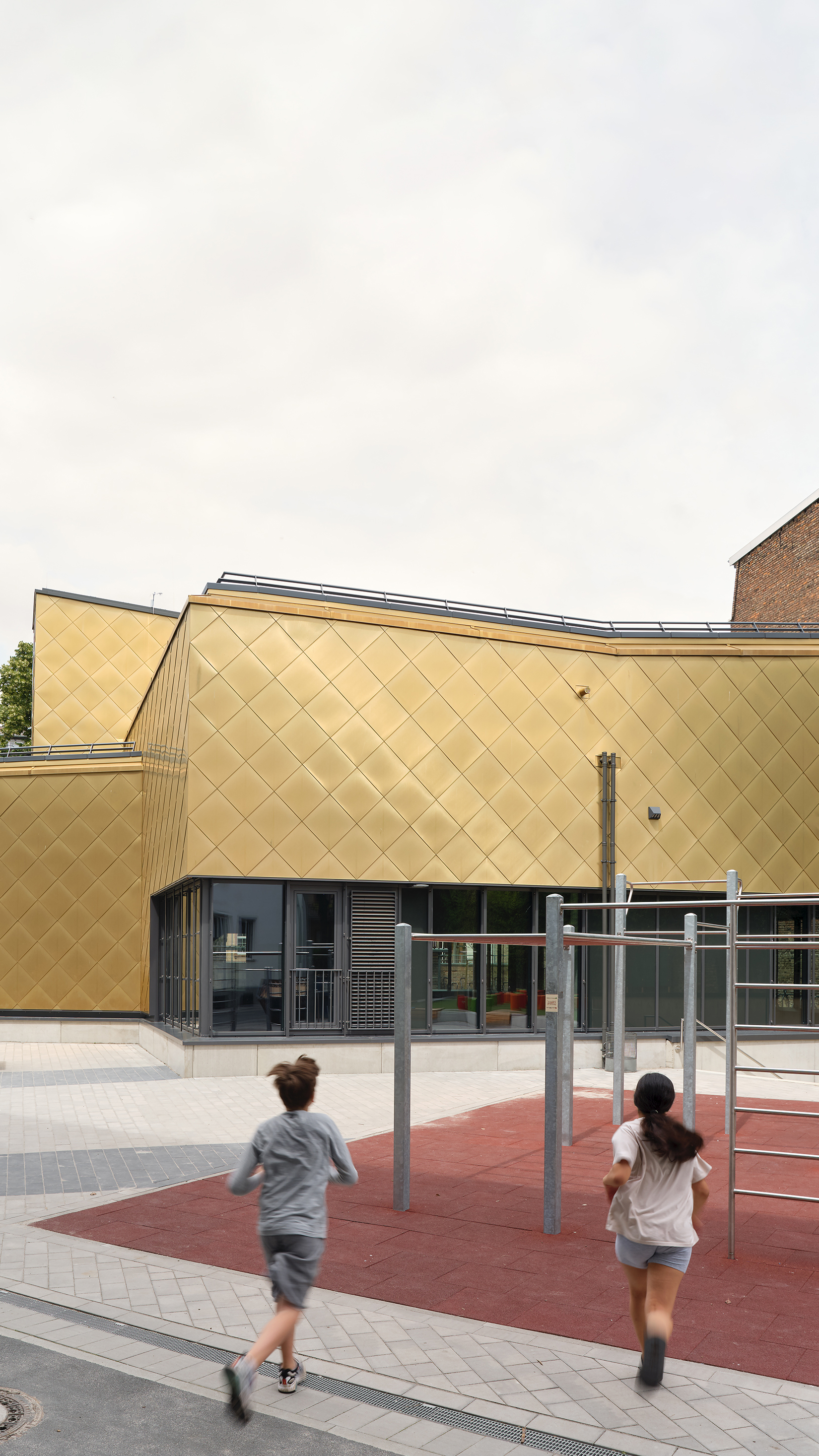 Kinder rennen über einen Spielplatz auf eine vielfach gestaffelten Sporthalle mit goldener Metallfassade zu.