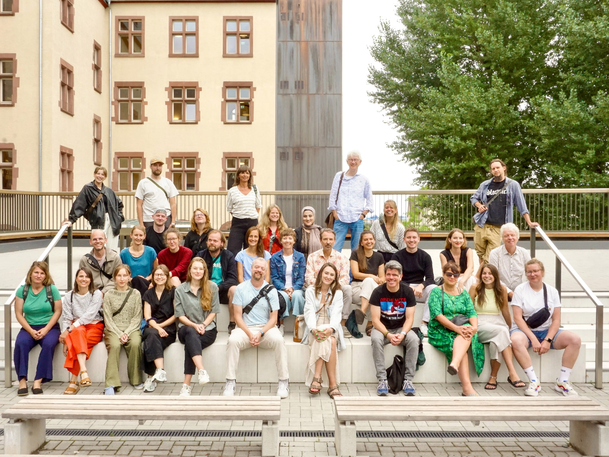 Eine Gruppe von erwachsenen Menschen sitzt und steht auf einer Sitzstufenanlage eines Schulhofs.