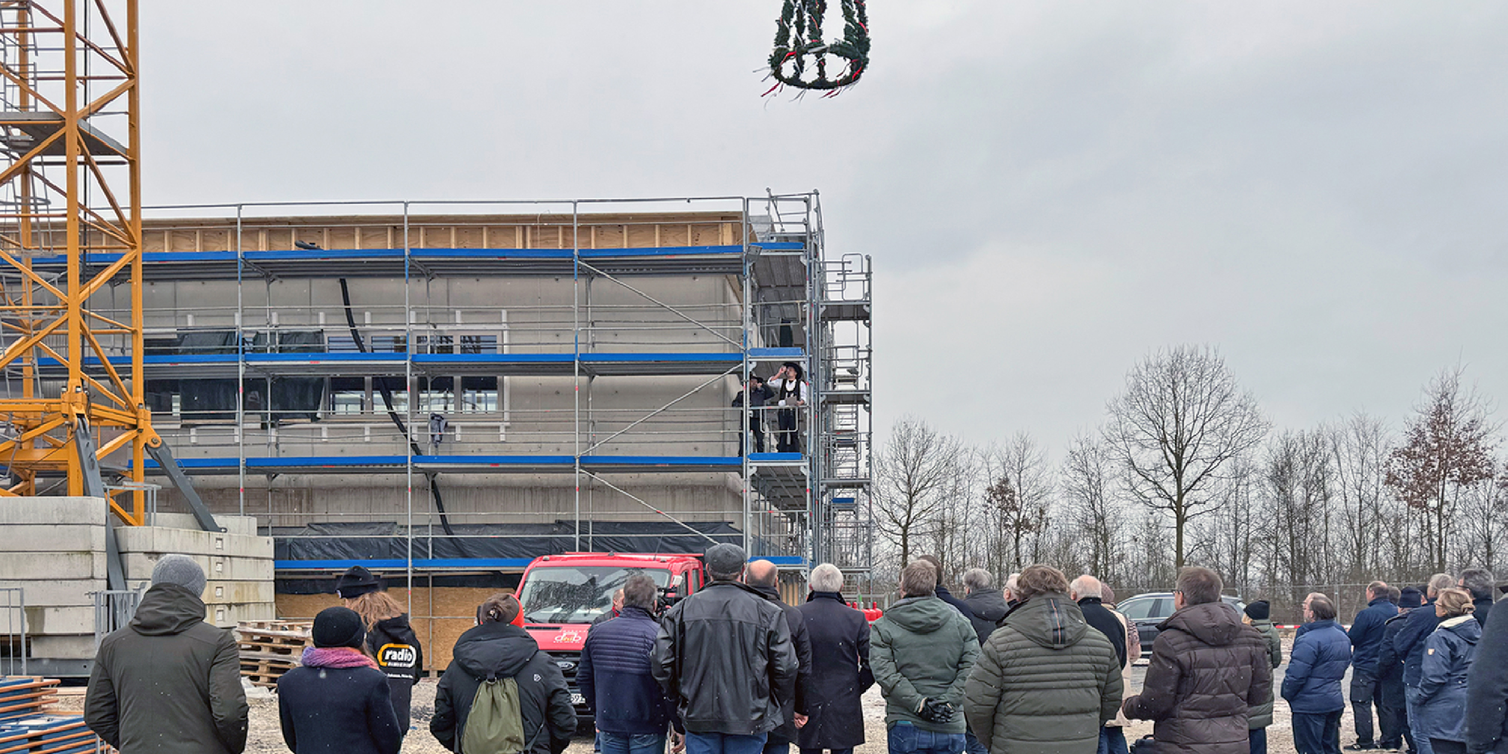 Eine Baustelle vor einem viergeschossigen Rohbau. Viele Menschen schauen zu einer Richtkrone und den Zimmerleuten hoch.