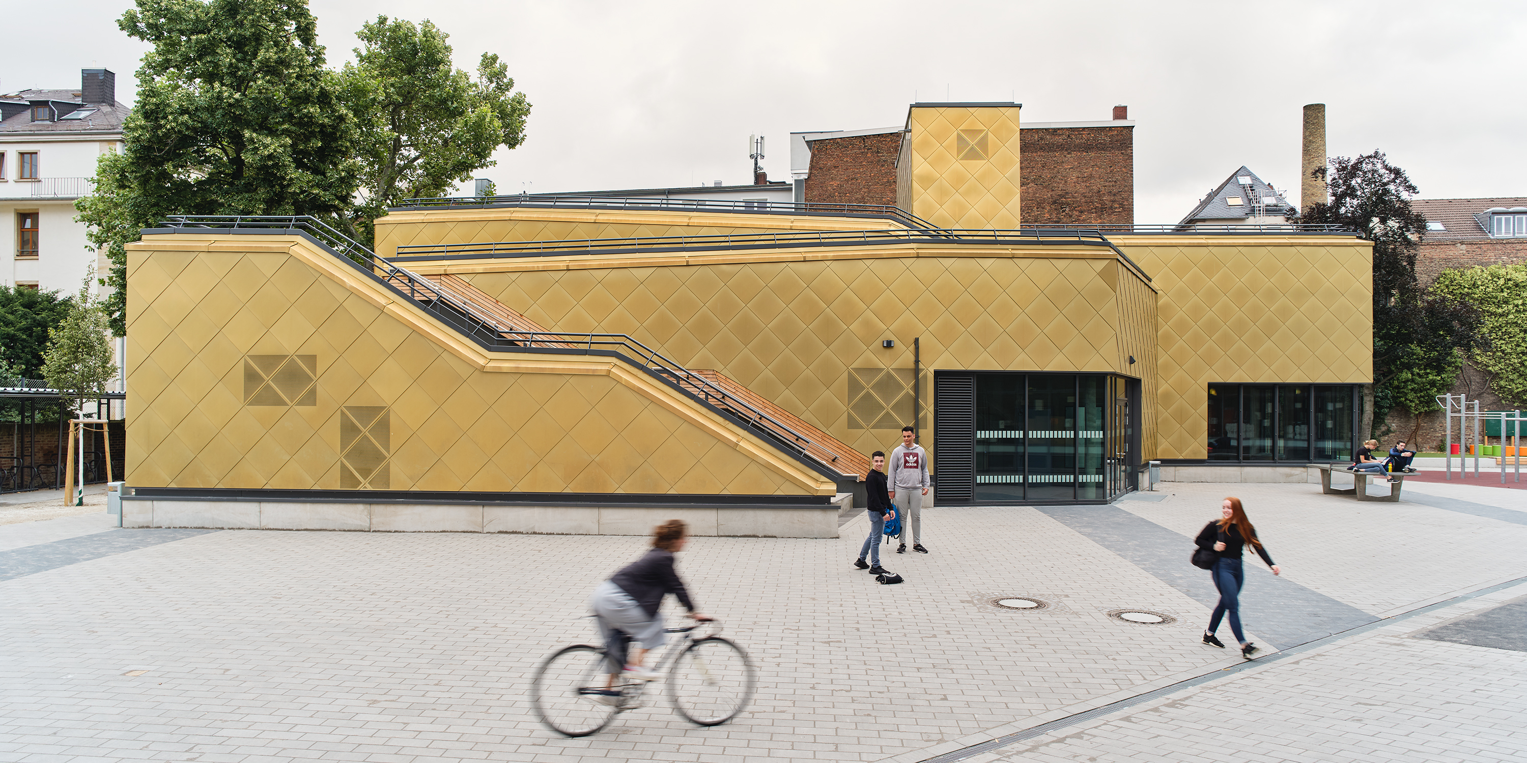 Schüler laufen oder fahren Fahrrad auf dem Schulhof vor dem Gebäude einer mehrfach gestaffelten Sporthalle. Die Fassade besteht aus goldenen Metalschindeln. Eine Treppe und Rampen führen vom Erdgeschoss auf das Dach.