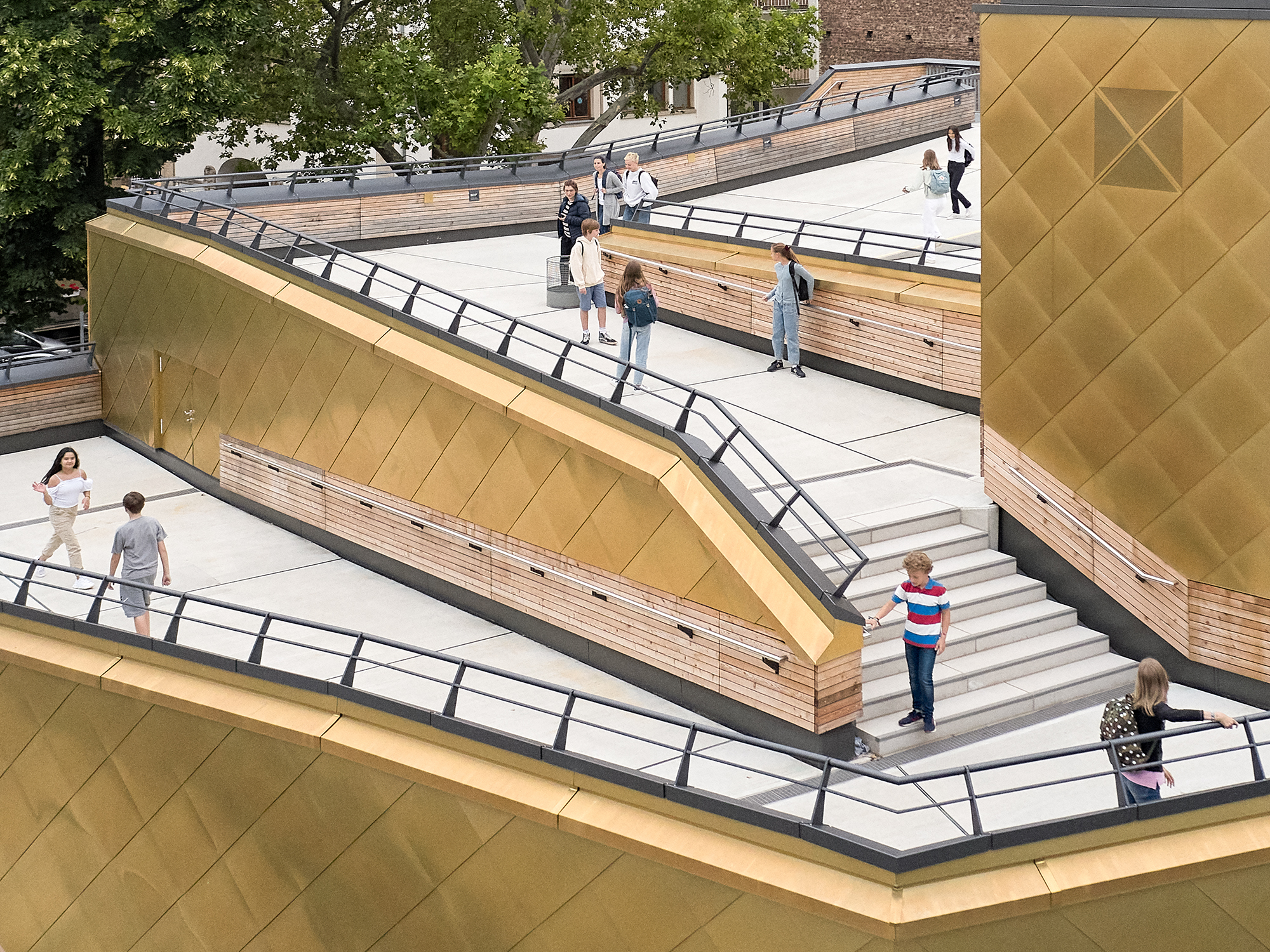 Schüler verteilen sich über Treppen und Rampen auf dem begehbaren Dach einer mehrfach gestaffelten Sporthalle. Die Fassade besteht aus goldenen Metalschindeln.