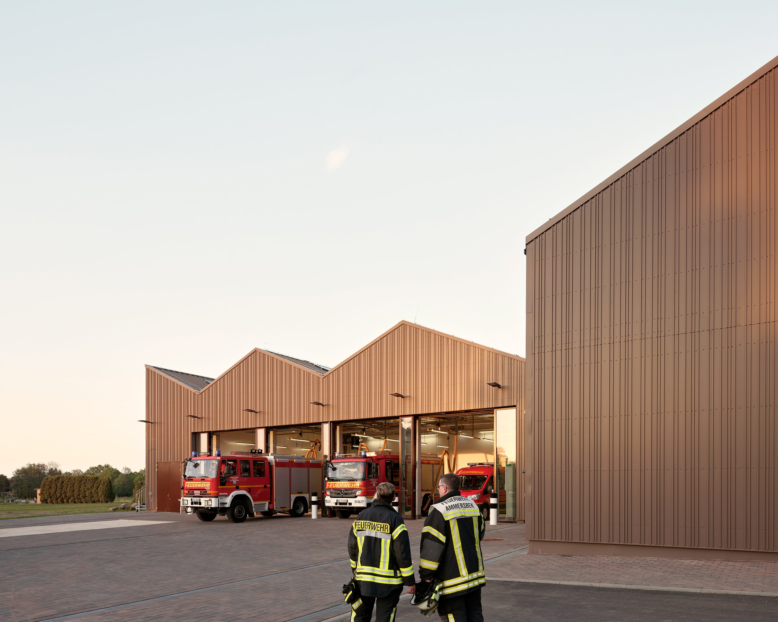 Zwei Feuerwehrleute vor einem Feuerwehrhauses mit geöffneten Toren zur Fahrzeughalle, in der Einsatzfahrzeuge stehen. Die Fassade ist aus rotbraunem Metall.