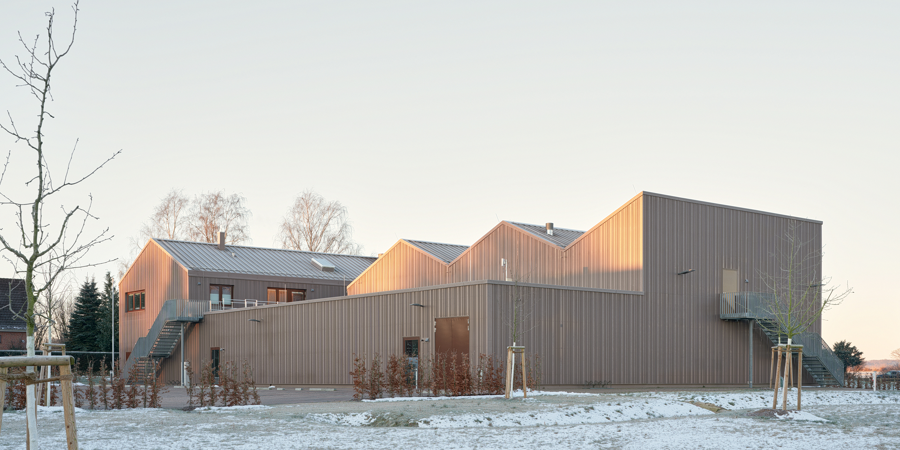 Winterliche Außenaufnahme einer Feuerwache mit rotbrauner Metallfassade. Mehrere Satteldächer strukturieren das Gebäude.
