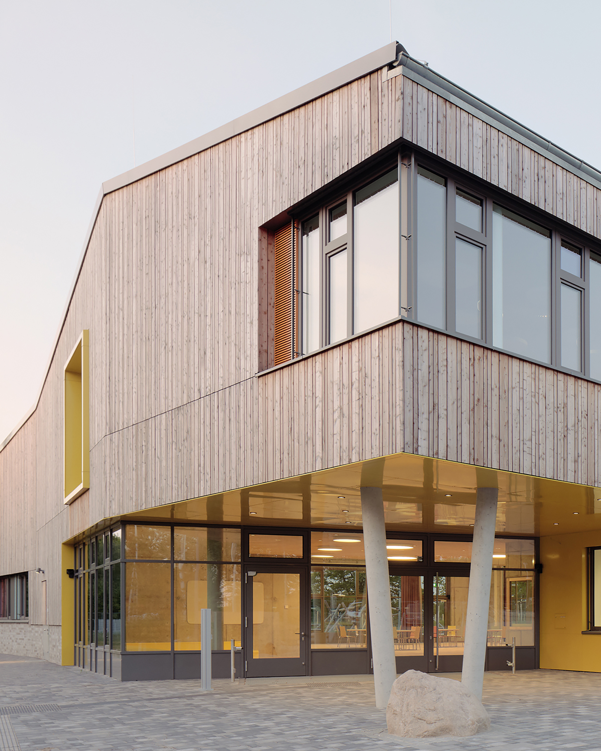 Außenaufnahme einer zweigeschossigen Schulgebäudes mit Holzfassade und und großen Fenstern. Eine Ecke des Obergeschoss kragt aus und wird von zwei schräg gestellten Stützen getragen, kräftige Farben betonen Fenster und Eingangsbereich.