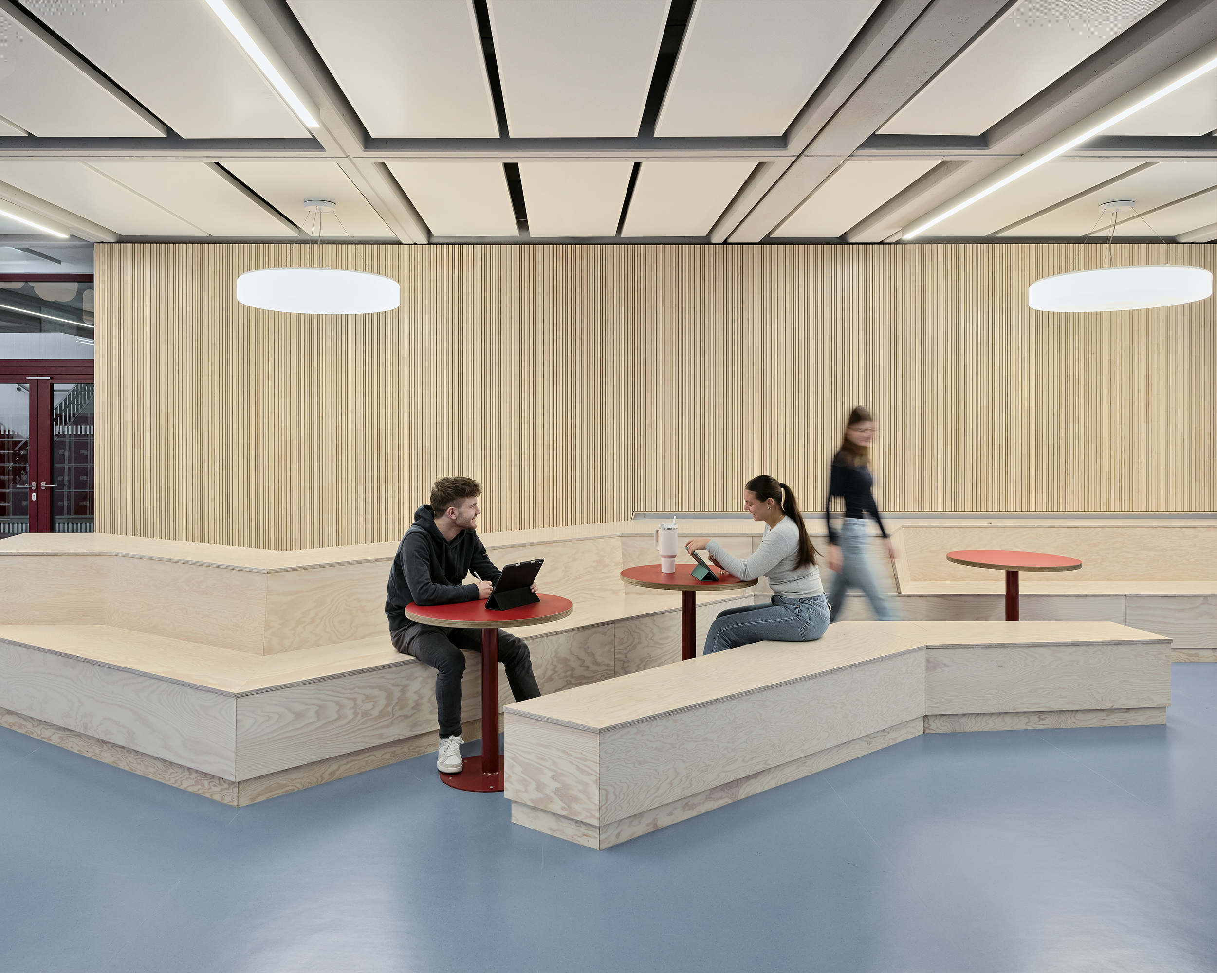 Schüler sitzen in der Lernlandschaft einer Schule auf fest eingebauten Möbeln mit runden Tischen. die Möbel und die Rückwand sind aus Holz, der Fußboden ist aus blauem Linoleum.