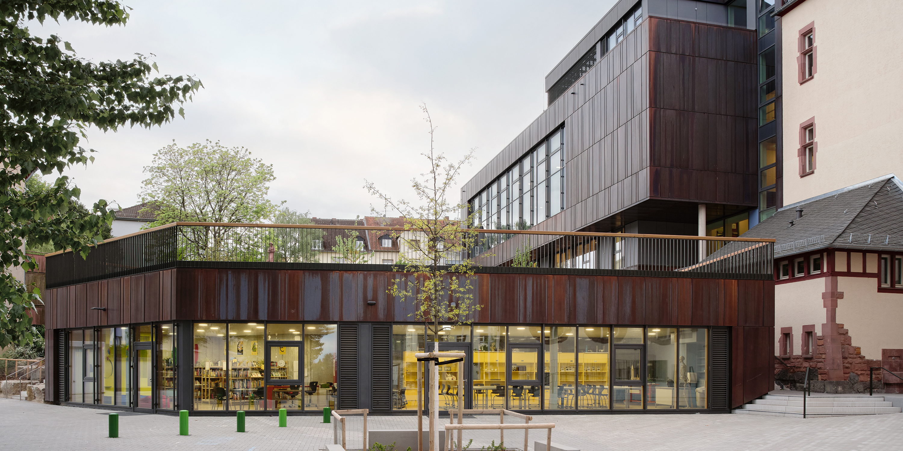 Ein Schulneubau mit dunkler Metallfassade und Schulhof. Aus einer großen Galsfassade schein warmes Licht