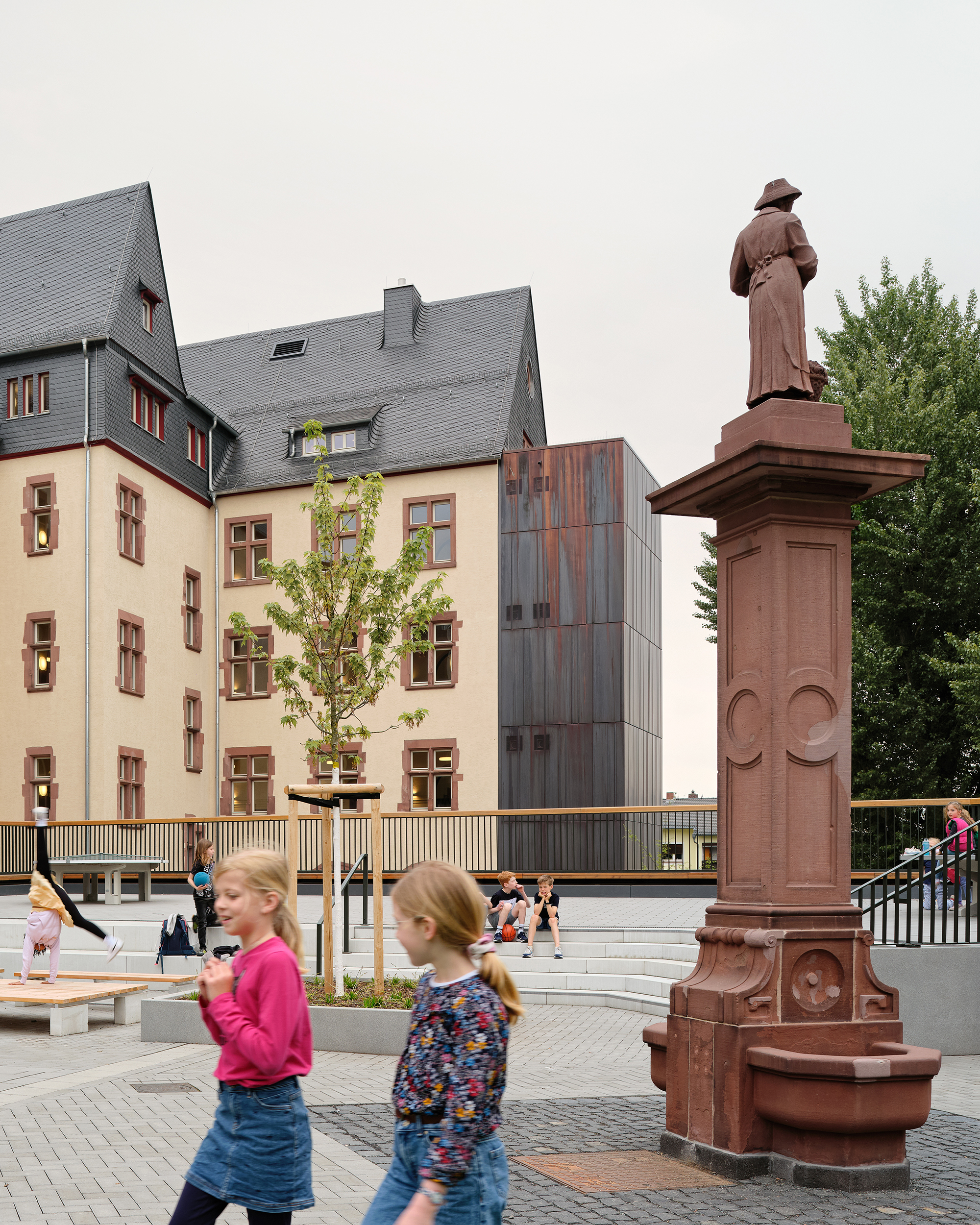 Zwei Kinder auf einem Schulhof vor einer historischen Brunnenskultur. Im Hintergrund ist ein historisches Schulgebäude