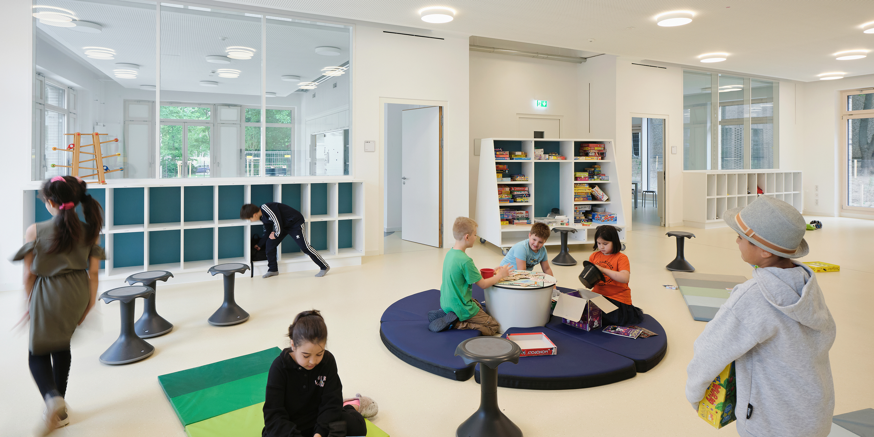Kinder spielen im großzügigen offen gestalteten Spiel- und Lernbereich in einem Cluster einer Grundschule. Teppiche, Matratzen und mobile Regale definieren den Raum.
