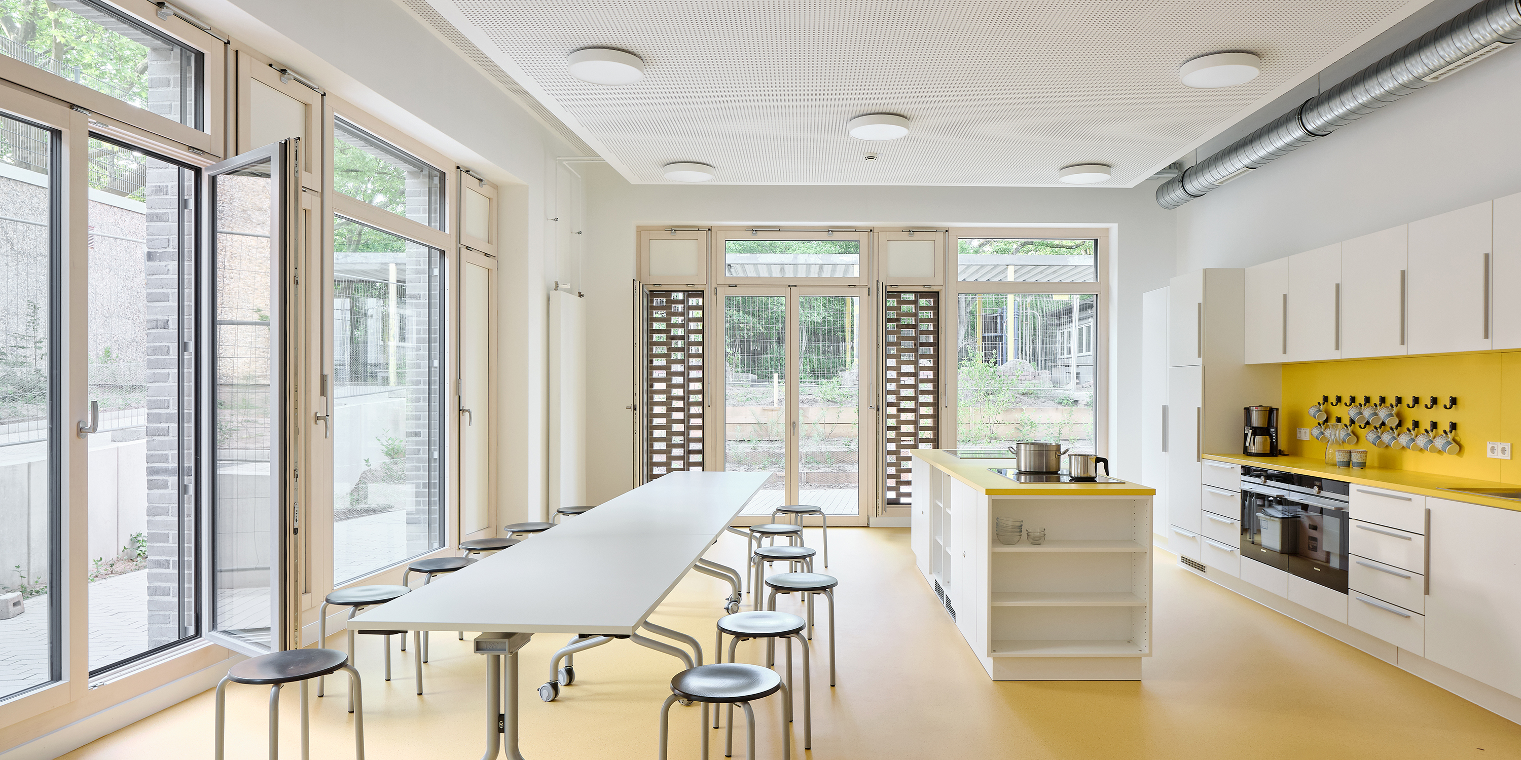 Eine Küche für Schulkinder. Zweisseitig sind bodentiefe Fenster, einige mit Wetterschutzgitter.