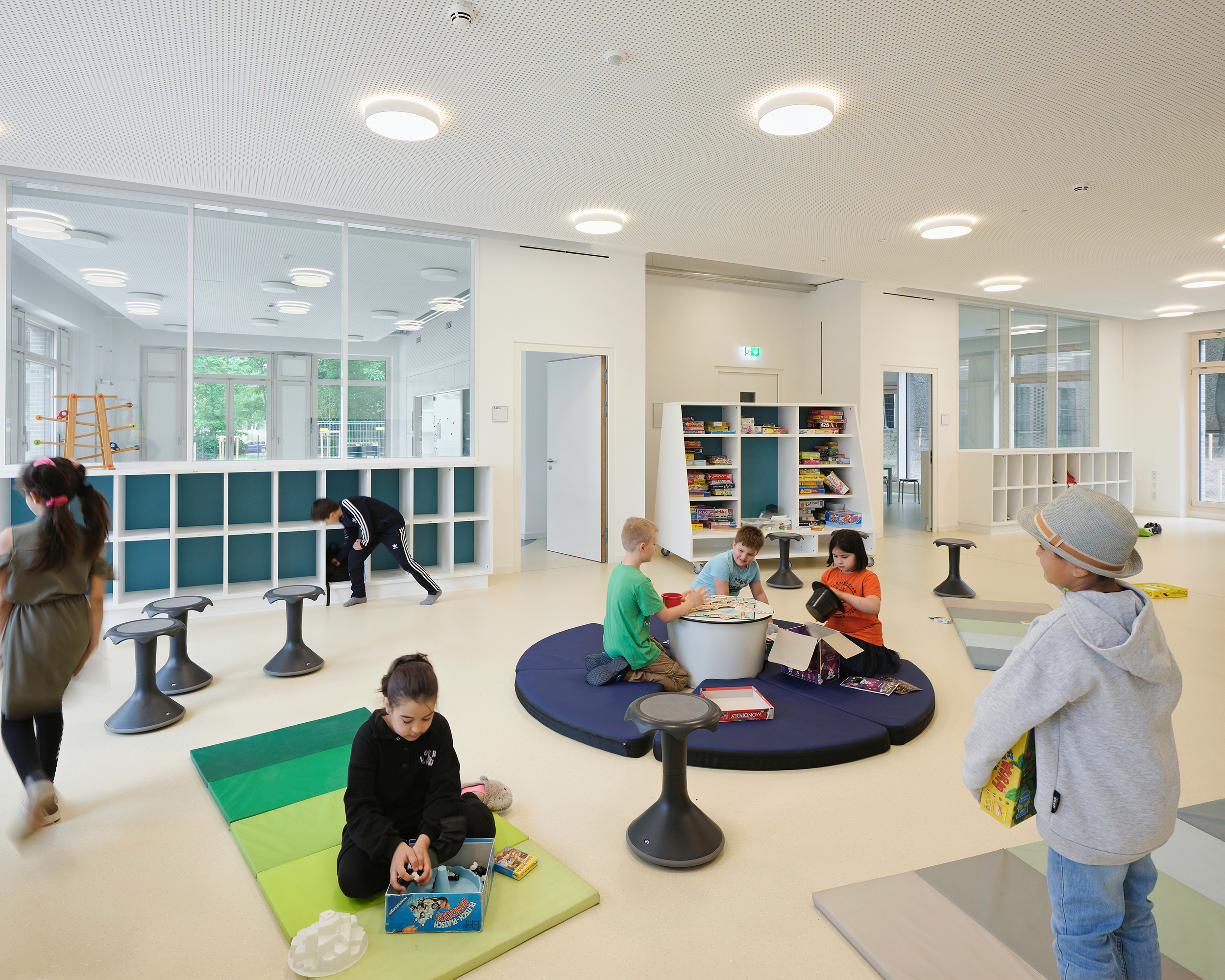 Kinder spielen im großzügigen offen gestalteten Spiel- und Lernbereich in einem Cluster einer Grundschule. Teppiche, Matratzen und mobile Regale definieren den Raum.